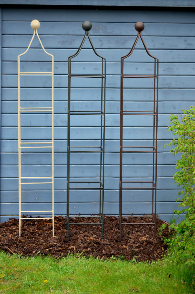 Obelisken Catania klein in pulverbeschichtet Sonderfarben RAL 8017 Schokobraun, RAL 6009 Tannengrün und RAL 9001 Cremeweiß aufgereiht in einem Beet Obelisken Catania klein in pulverbeschichtet Sonderfarben RAL 8017 Schokobraun, RAL 6009 Tannengrün und RAL 9001 Cremeweiß aufgereiht in einem Beet